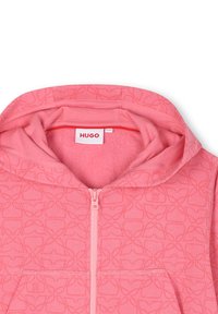Haut à capuche zippé rose avec un motif floral subtil, texture douce et capuche. Comprend une étiquette logo à l'intérieur du col et des poches avant.