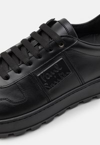 Basket en cuir noir avec des lacets noirs épais, un patch logo rectangulaire en relief et une semelle solide, vue de côté en gros plan.