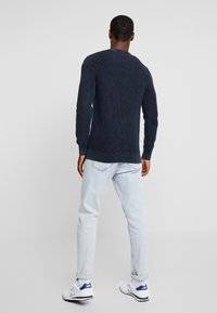 Marineblaue gerippte Strickjacke mit langen Ärmeln, Rundhalsausschnitt und taillierter Passform, kombiniert mit hellblauen Jeans und weißen Sneakers.