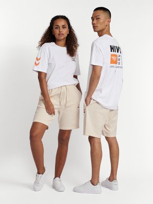 Vrouw en man staan in witte grafische T-shirts en beige shorts met witte sneakers tegen een effen achtergrond.