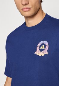 Marineblå bomulds t-shirt med grafisk design i pastelfarver og sky mønster omkring et centralt logo på venstre brystområde.