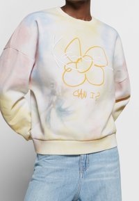 Persoon draagt een pastel tie-dye sweater met gele bloem en witte geborduurde tekst, gecombineerd met lichtblauwe spijkerbroek, handen achter de rug.