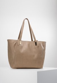 Beige nahkalaukku, jossa on kaksi pitkää kahvaa ja tyylikkäitä kultasävyisiä yksityiskohtia. Laukku on muotoilultaan jämäkkä ja sen pinta on sileä.