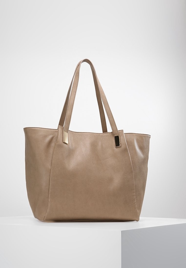 Beige nahkalaukku, jossa on kaksi pitkää kahvaa ja tyylikkäitä kultasävyisiä yksityiskohtia. Laukku on muotoilultaan jämäkkä ja sen pinta on sileä.