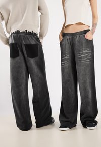 Schwarze, weit geschnittene Hose mit texturiertem Finish, ausgestattet mit zwei vorderen Taschen und zwei schwarzen Stofftaschen auf der Rückseite. Abgenutztes Detail am Bund.