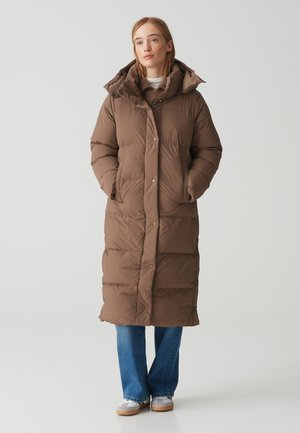 Long brown puffer coat with a hood, featuring a button closure, quilted texture, and side pockets, worn over blue jeans and sneakers.