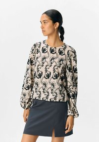 Blouse à manches longues en crème avec broderie florale noire, présentant un col rond et des manches froncées. Associée à une jupe gris foncé.
