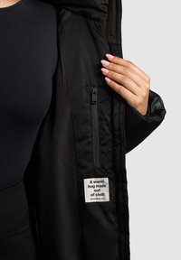 Schwarze isolierte Jacke mit glatter Außenseite und weichem Innenfutter. Verfügt über eine Reißverschlusstasche und ein Stoffetikett mit dem Text: "Eine warme Umarmung aus Stoff."