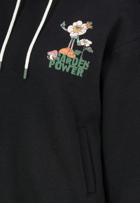 Zwarte sweater met een grafisch ontwerp van bloemen, paddenstoelen en een karakter. Kap met witte trekkoorden en groene accenten.