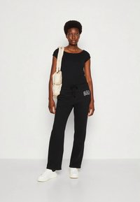 Žena v čiernom tričku a teplákoch s logom GAP, v bielych teniskách, s béžovou kabelkou na ramene, stojaca proti jednoduchému svetlému pozadiu.