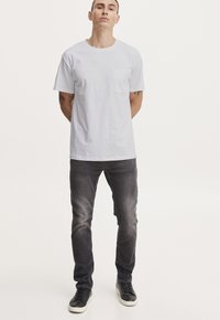 Camiseta blanca de manga corta con un bolsillo en el pecho, combinada con jeans gris oscuro y zapatillas negras, de pie contra un fondo claro y liso.