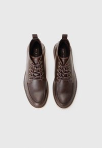 Botas de cuero marrón hasta el tobillo con punta redonda, costuras mínimas y ojales dorados para los cordones. Con una lengüeta para facilitar el uso.