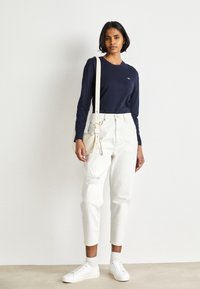 Marineblå langermet topp, hvite høytlivs bukser med slitte detaljer, hvite sneakers og en lys crossover-veske. Minimalistisk design.