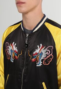 Homme portant une veste bomber en satin noir et jaune avec des motifs de dragons colorés brodés sur la poitrine et un col rayé.