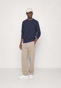 Sweatshirt azul-marinho com um pequeno logo em patch, calças largas bege e ténis brancos. A pessoa usa um boné de cor clara.