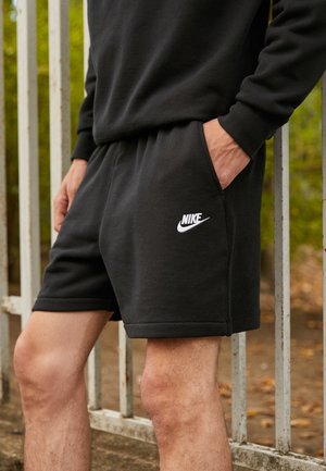 Person trägt schwarze Nike-Shorts, hat eine Hand in der Tasche und steht draußen in der Nähe eines Metallzauns, im Hintergrund ist Grün zu sehen.