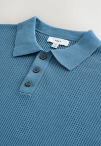 Polo de rayas azules con un cuello suave, presenta una tapeta de tres botones y botones oscuros. Etiqueta visible con información de la marca y el tamaño.