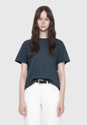 Young woman with long brown hair wearing a dark blue t-shirt tucked into white pants with a black belt, standing against a plain light background.
