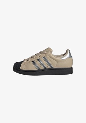 Beige leren sneaker met drie zilveren strepen, een zwarte rubberen neus en een zwarte zool. Bevat zilveren accenten op de hak en veter.