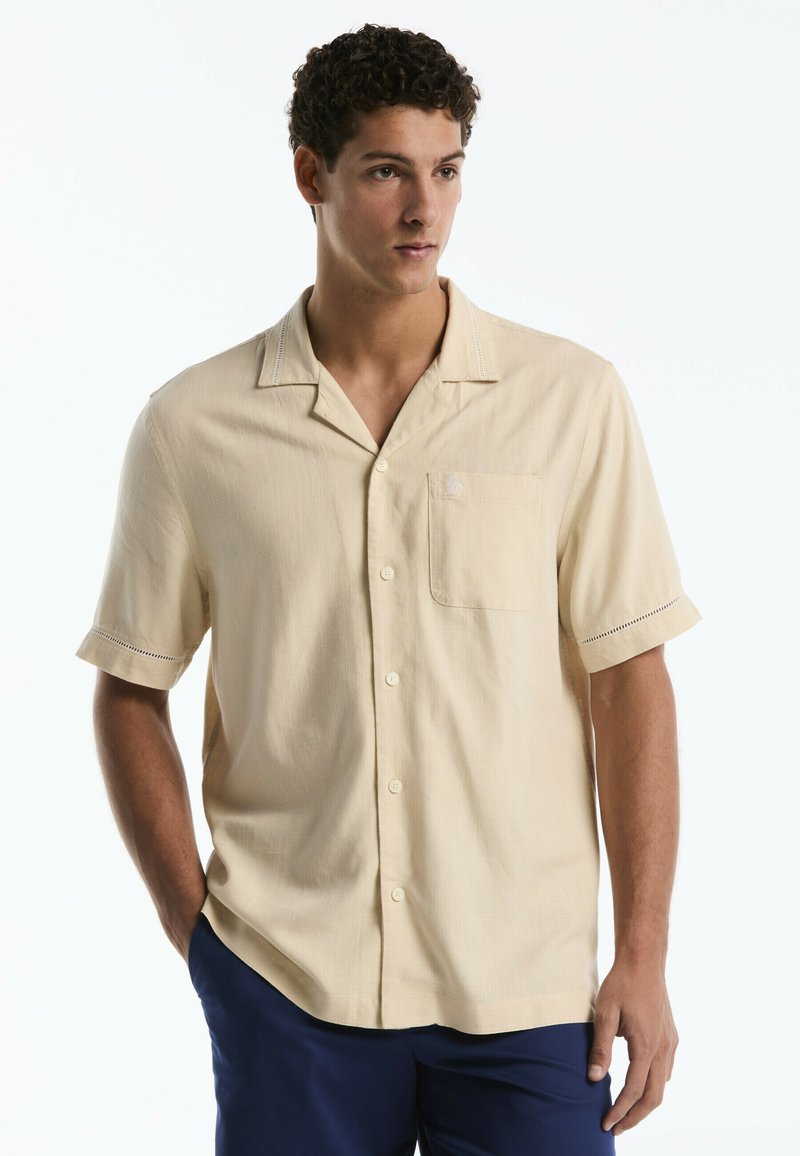 Man with short curly hair wearing a beige short-sleeve button-up shirt and navy blue pants, standing with one hand in pocket.