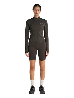 Frau, die nach vorne schaut, trägt eine dunkle langärmelige Reißverschlussjacke mit „HALO“-Logo, passende Radlerhosen, weiße Socken und schwarze Sneakers.