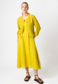 Kollane kudunud kardigan tekstuursete mullidega, koos sobiva lina kleidiga, millel on ees olev lõhik. Pruunid rihmadega sandaalid täiustavad ilmet.