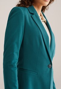 Turkizno moder suknjič iz gladkega blaga z zarezo na ovratniku, zapenjanjem na en gumb in stranskimi žepi za prilagojeno prileganje.