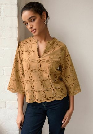 Blusa beige con scollatura a V e ampie maniche a 3/4, caratterizzata da motivi intagliati decorativi e bordi smerlati. Abbinata a jeans blu scuro.