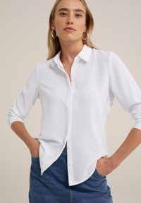 Camisa blanca de manga larga con botones y cuello, con plantilla frontal y dobladillo redondeado; combinada con una falda de mezclilla. Tejido suave, textura lisa.