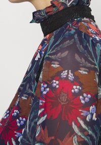 Vícebarevná květinová blůza s řaseným límcem, průsvitnou tkaninou a černým texturovaným páskem kolem výstřihu. Obsahuje výrazné červené a modré vzory.