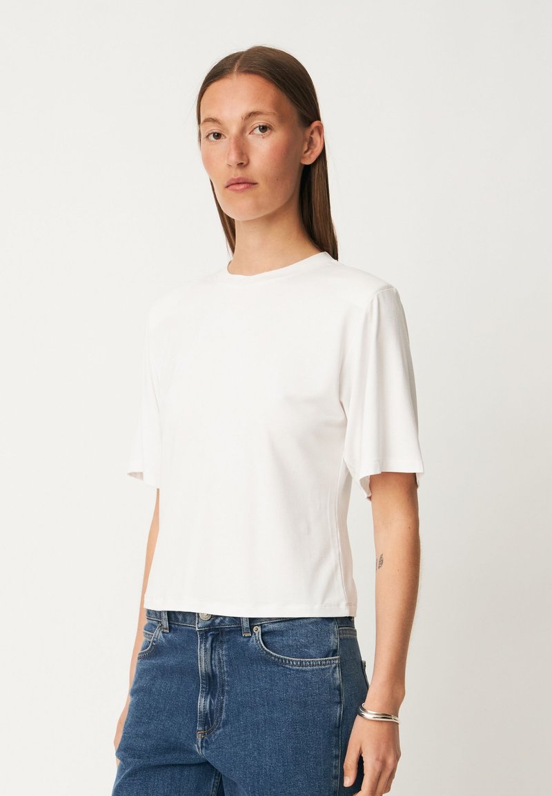 Femme aux cheveux bruns raides, portant un haut blanc uni à manches courtes et un jean bleu, debout devant un fond clair.