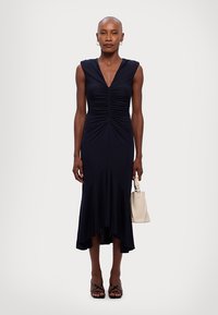 Navy sleeveless dress with gathered bodice, flared hem, and asymmetrical cut. Paired with a light handbag and strappy black heels.