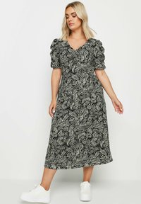 Svartvit blommig maxiklänning med korta puffärmar och en djup V-ringning, i kombination med vita sneakers på en neutral bakgrund.