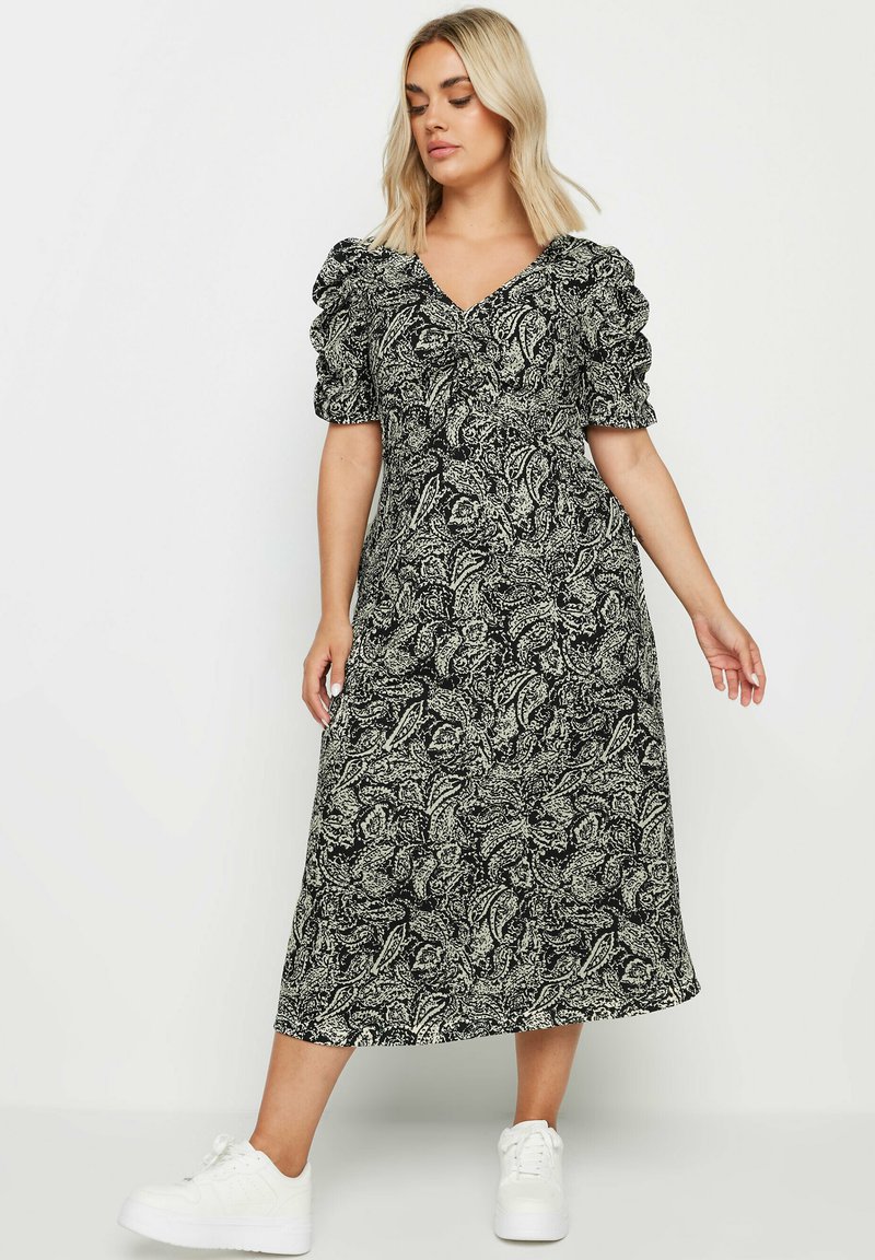 Svartvit blommig maxiklänning med korta puffärmar och en djup V-ringning, i kombination med vita sneakers på en neutral bakgrund.