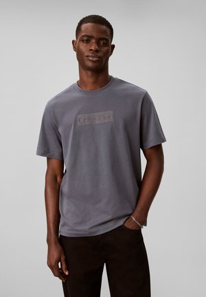 Jeune homme portant un t-shirt gris Calvin Klein à manches courtes, debout devant un fond clair uni, avec une main dans la poche.