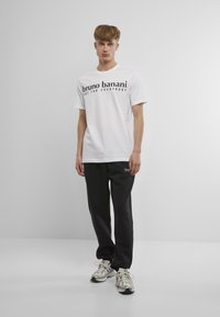 Bílá bavlněná tričko s černým textovým logem "bruno banani" a "not for everybody"; v kombinaci s černými tepláky se malým logem.