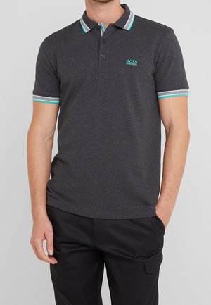 Man draagt een donkergrijze Hugo Boss polo met een turkooise en witte gestreepte kraag en mouwranden, gecombineerd met een zwarte broek.