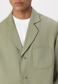 Light olive green jacket with a notched collar, two buttons, and a chest pocket. Smooth fabric with a subtle texture. Worn over a white shirt.