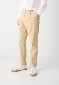 Mężczyzna ubrany w beżowe spodnie o luźnym kroju i białe trampki z gumową podeszwą, stojący z jedną ręką w kieszeni na jednolitym tle.