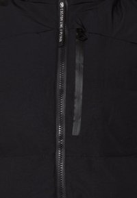 Sort jakke med lynlås i fuld længde og en ekstra lomme med lynlås. Materialet virker let med en glat tekstur.