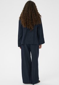 Žena s dlhými kučeravými vlasmi oblečená v tmavomodrom širokých nohaviciach, stojaca chrbtom k jednoliatemu svetlému pozadiu.