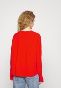 Roter Strickpullover mit lockerer Passform, Rundhalsausschnitt und langen Ärmeln, kombiniert mit hellblauen Jeans. Flache Textur, minimale Details.