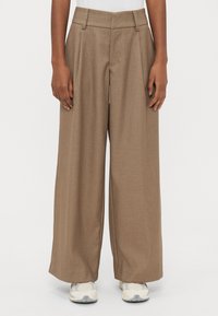 Bézs krāsas plašu kāju bikses ar pleatiem, izgatavotas no teksturēta auduma. Tām ir augsta josta un kabatas, kombinētas ar gaišiem sporta apaviem.