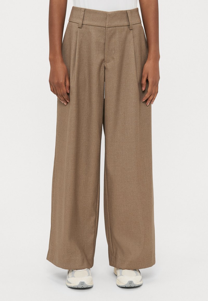 Bézs krāsas plašu kāju bikses ar pleatiem, izgatavotas no teksturēta auduma. Tām ir augsta josta un kabatas, kombinētas ar gaišiem sporta apaviem.