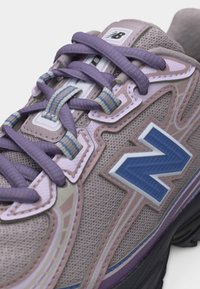 Ανοιχτό μωβ και μπεζ αθλητικό παπούτσι New Balance με μπλε λογότυπο "N", μωβ κορδόνια και λεπτομέρειες από δίχτυ στο πάνω μέρος και μαύρη σόλα.