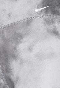 Legíny Nike v šedém batikovaném vzoru s hladkou texturou. Mají bílou logo a viditelné detaily švů.