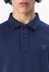 Polo azul marino de algodón, con un cuello clásico, dos botones negros y un pequeño logotipo de un árbol bordado en el pecho.