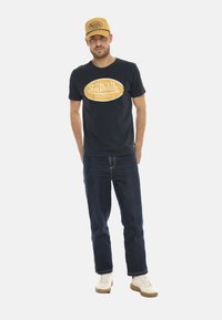 T-shirt noir avec logo "Von Dutch" jaune, jean sombre et baskets beiges. Une casquette dorée avec des détails de logo vient compléter le look.