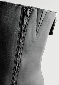 Stivale in pelle nera con cerniera laterale, dotato di dettagli metallici e superficie texture. Il design unico include fessure verticali nella parte superiore.