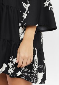 Schwarzes Kleid mit weißen Blumenmustern, gestuften Rüschen am Saum und einem fließenden Design. Glattes Material, kurze Ärmel und eine lockere Passform.